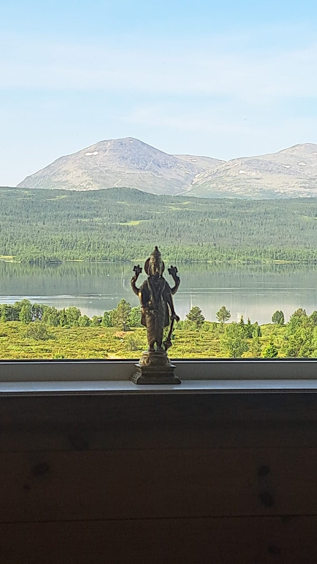 Small bronze deity statue on a windowsill overlooking a Norwegian lake and mountains