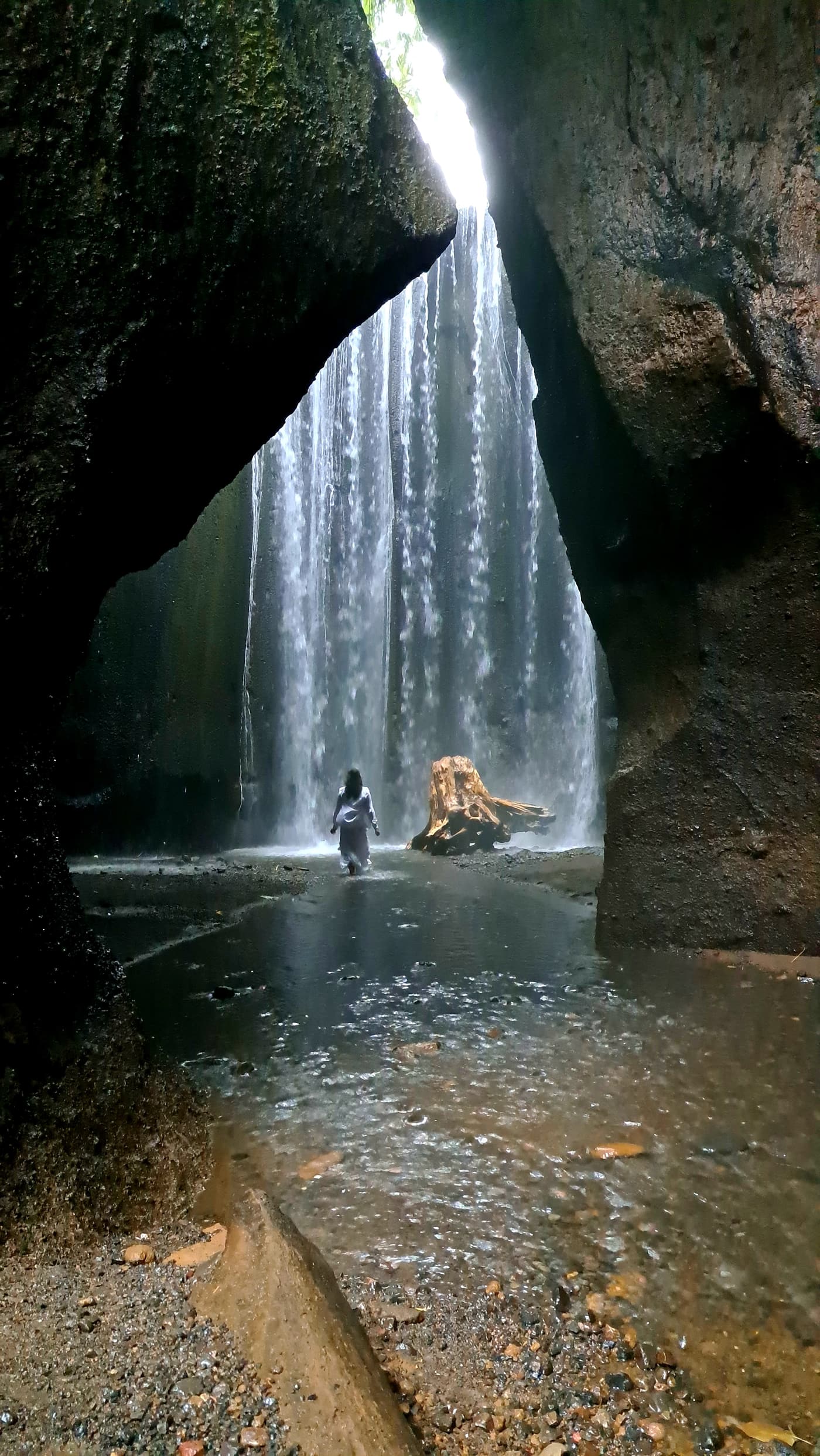 Boyana Vancheva in a white dress walking towards a curtain of falling water inside a Balinese rock canyon