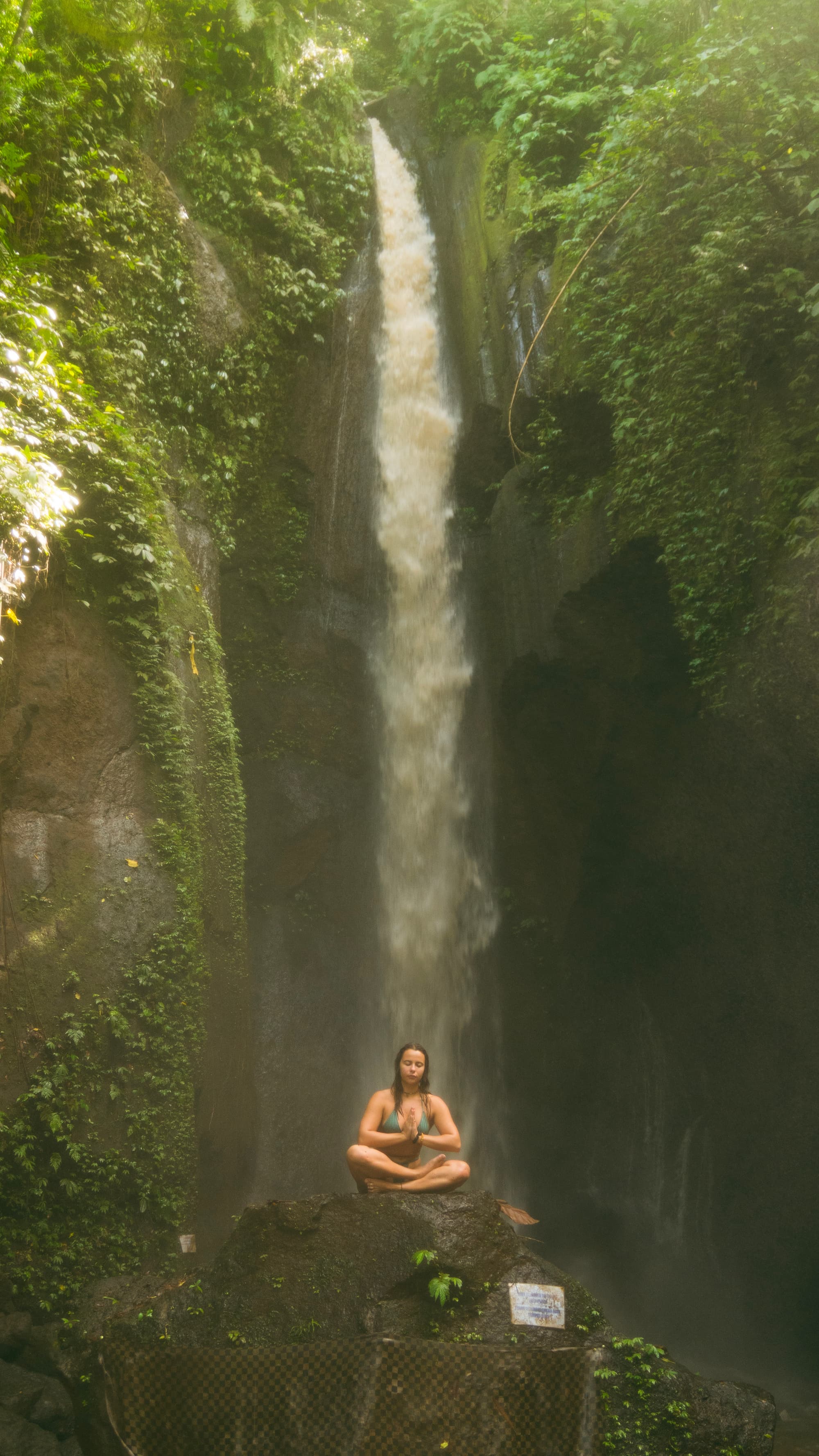 Boyana Vancheva seated in lotus meditation on a rock beneath a tall jungle waterfall in Bali