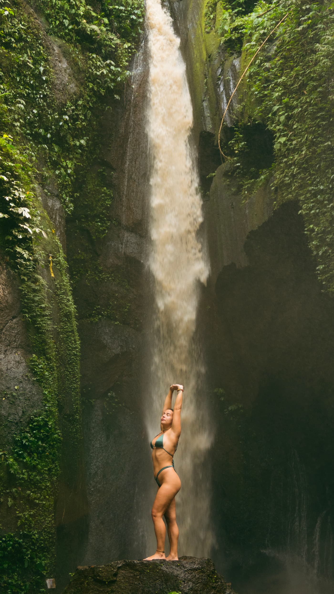 Boyana Vancheva standing with arms raised overhead on a rock beneath a tall Balinese waterfall